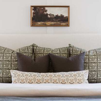 Framed landscape painting with golden-brown border hanging above bed with patterned headboard and decorative pillows.