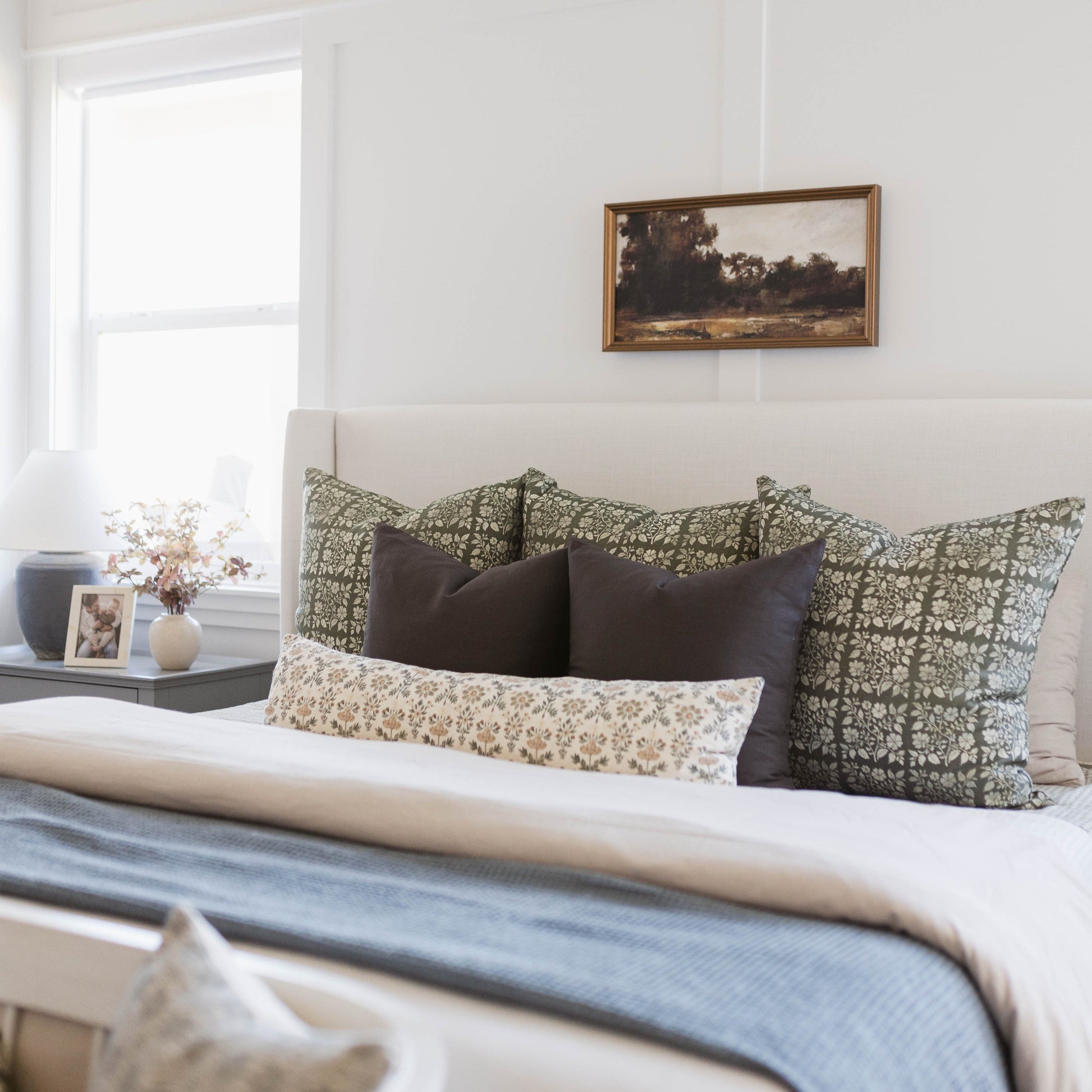 Framed landscape artwork with brown wooden frame hanging above bed with patterned pillows.