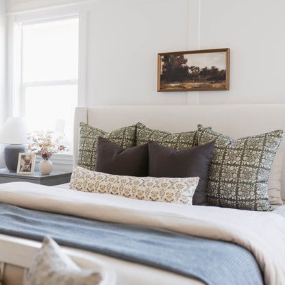 Framed landscape artwork with brown wooden frame hanging above bed with patterned pillows.
