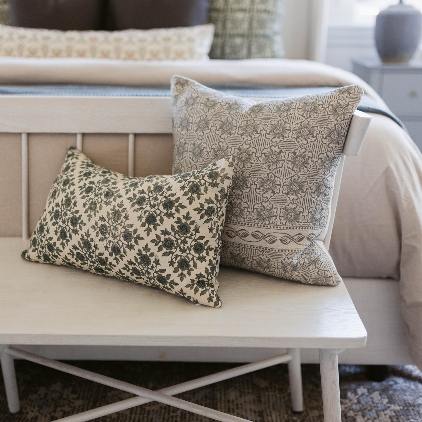 Two decorative pillows with damask and floral patterns in cream and sage tones displayed on a white wooden bench.