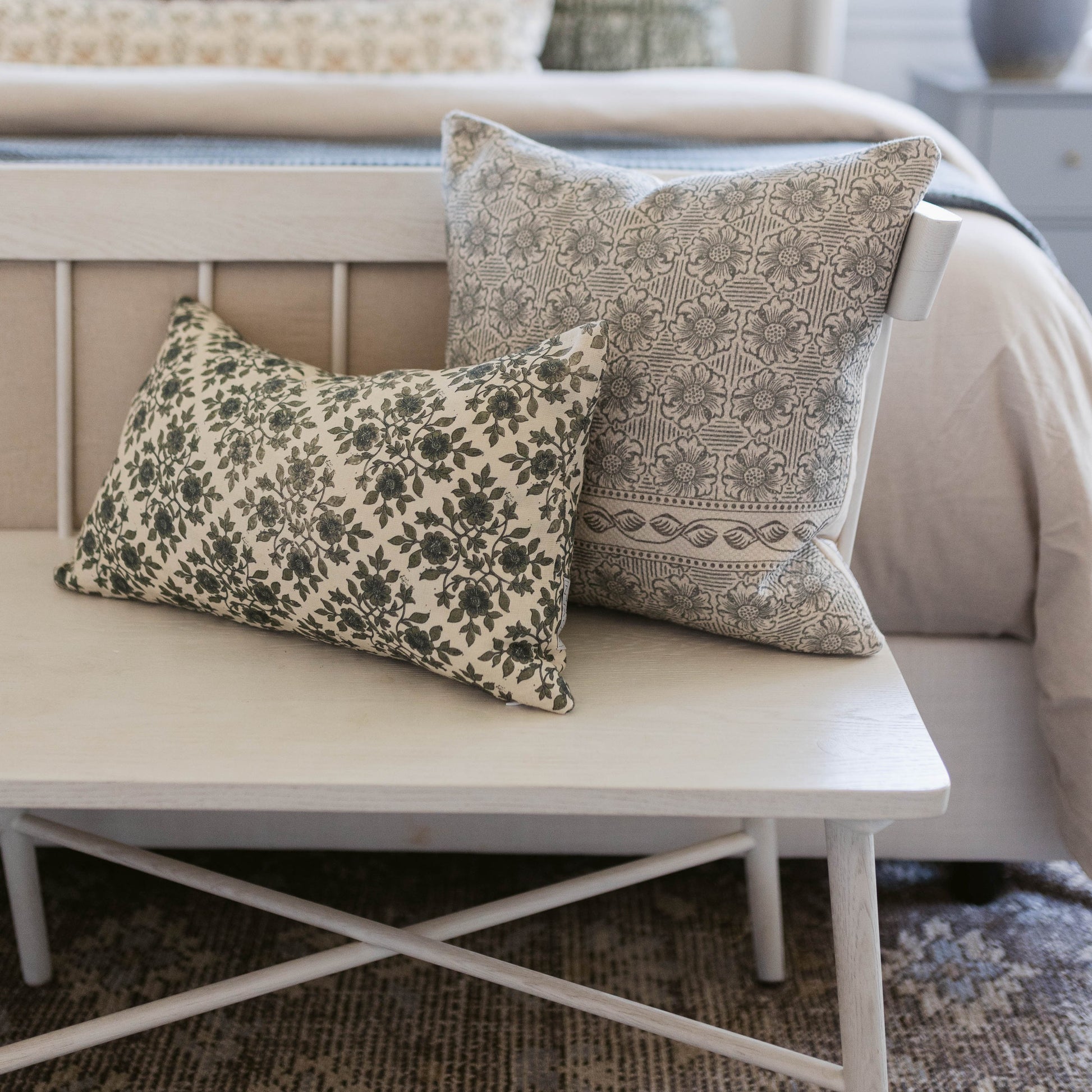 Two rectangular lumbar pillows with geometric and floral patterns in cream and sage green on a white bench.