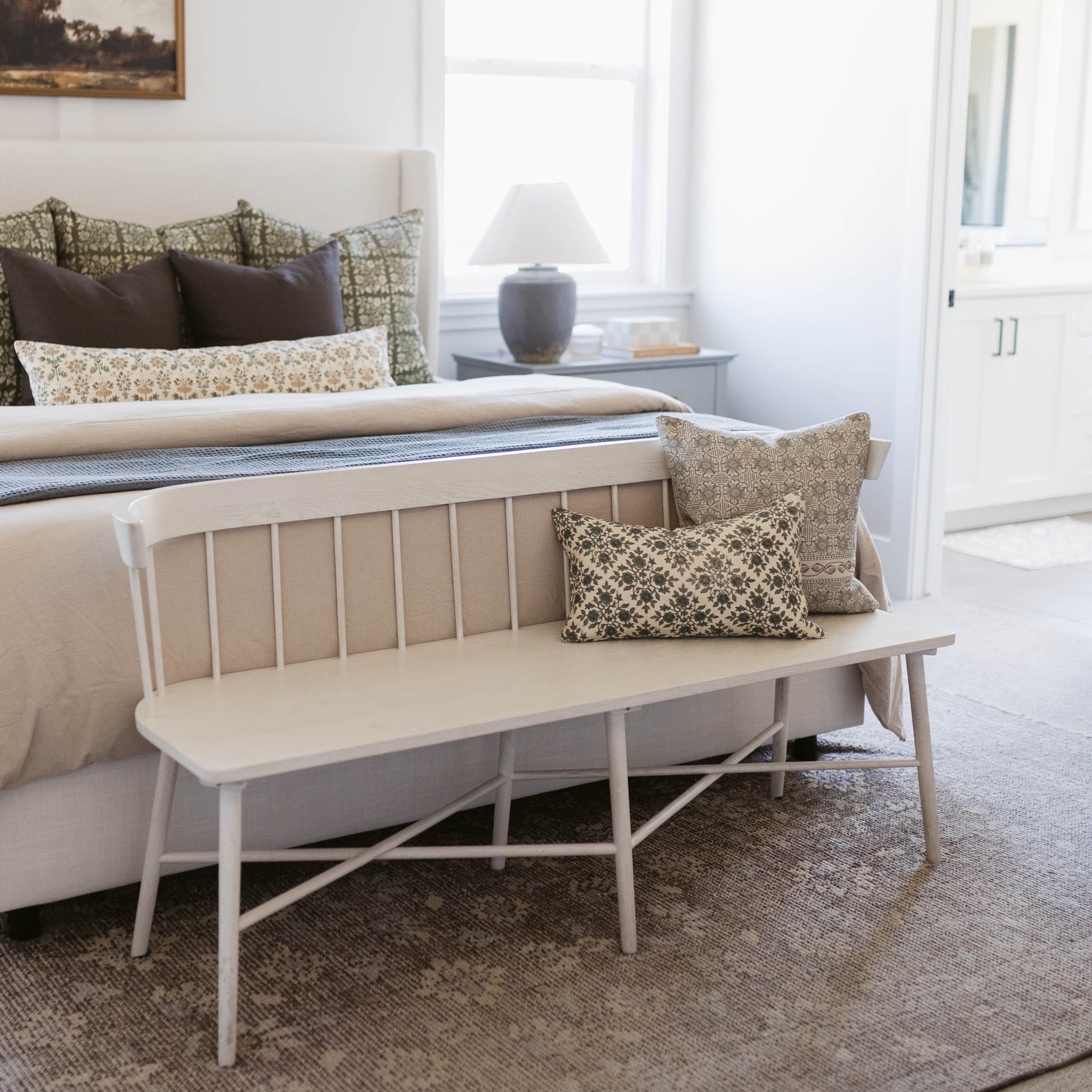 Tan and cream patterned lumbar pillow displayed on a white wooden bench at the foot of a bed.