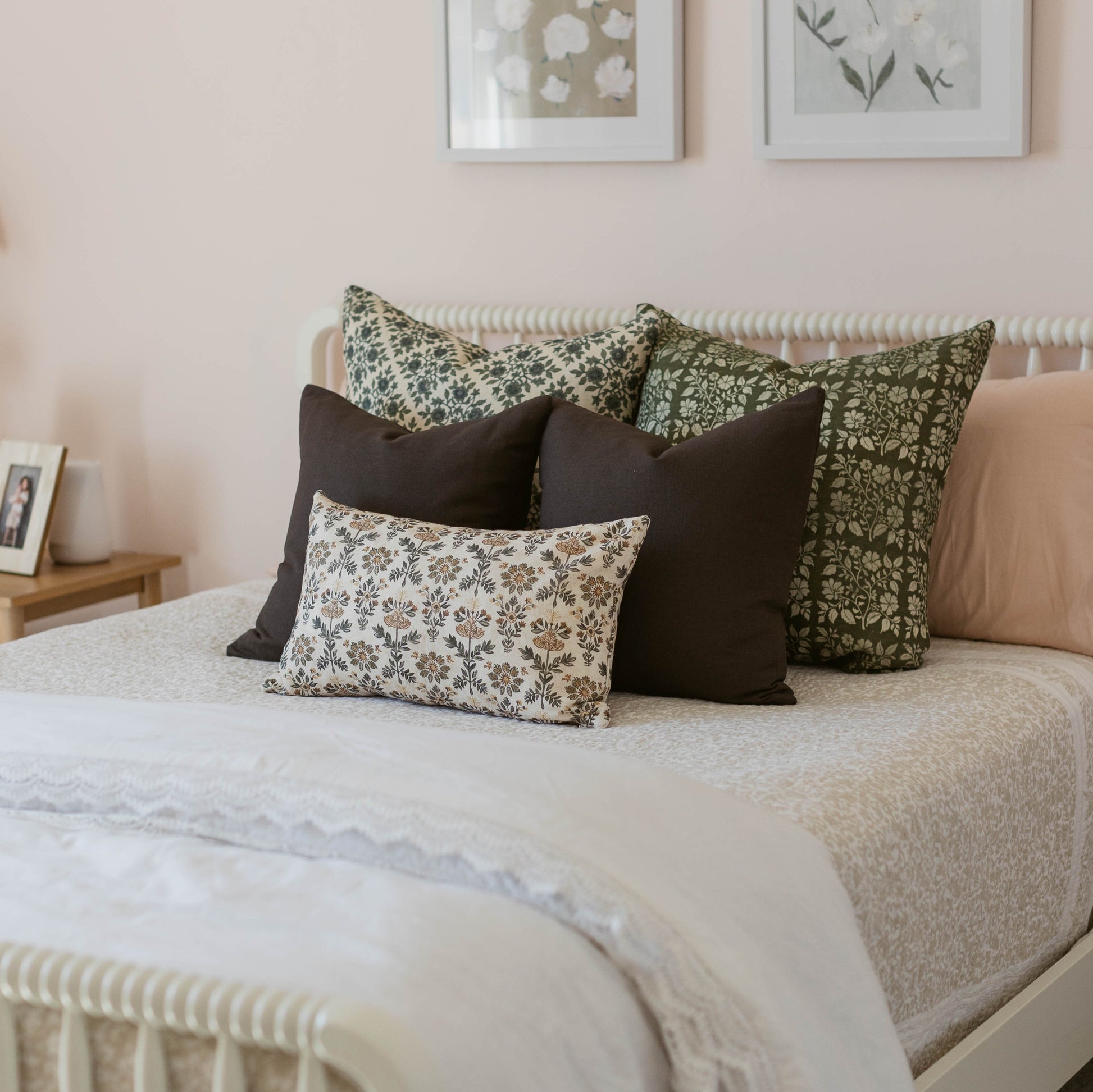 Linen pillow with small-scale evergreen floral print in cream and sage green, displayed on bed with solid black pillows.