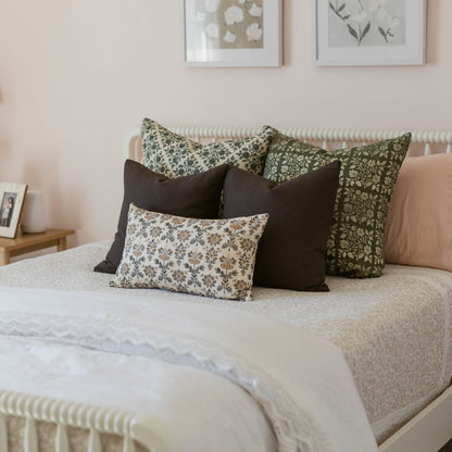 Linen pillow with small-scale evergreen floral print in cream and sage green, displayed on bed with solid black pillows.