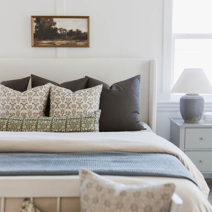 Framed landscape artwork with gold frame hanging above white upholstered bed with patterned pillows.