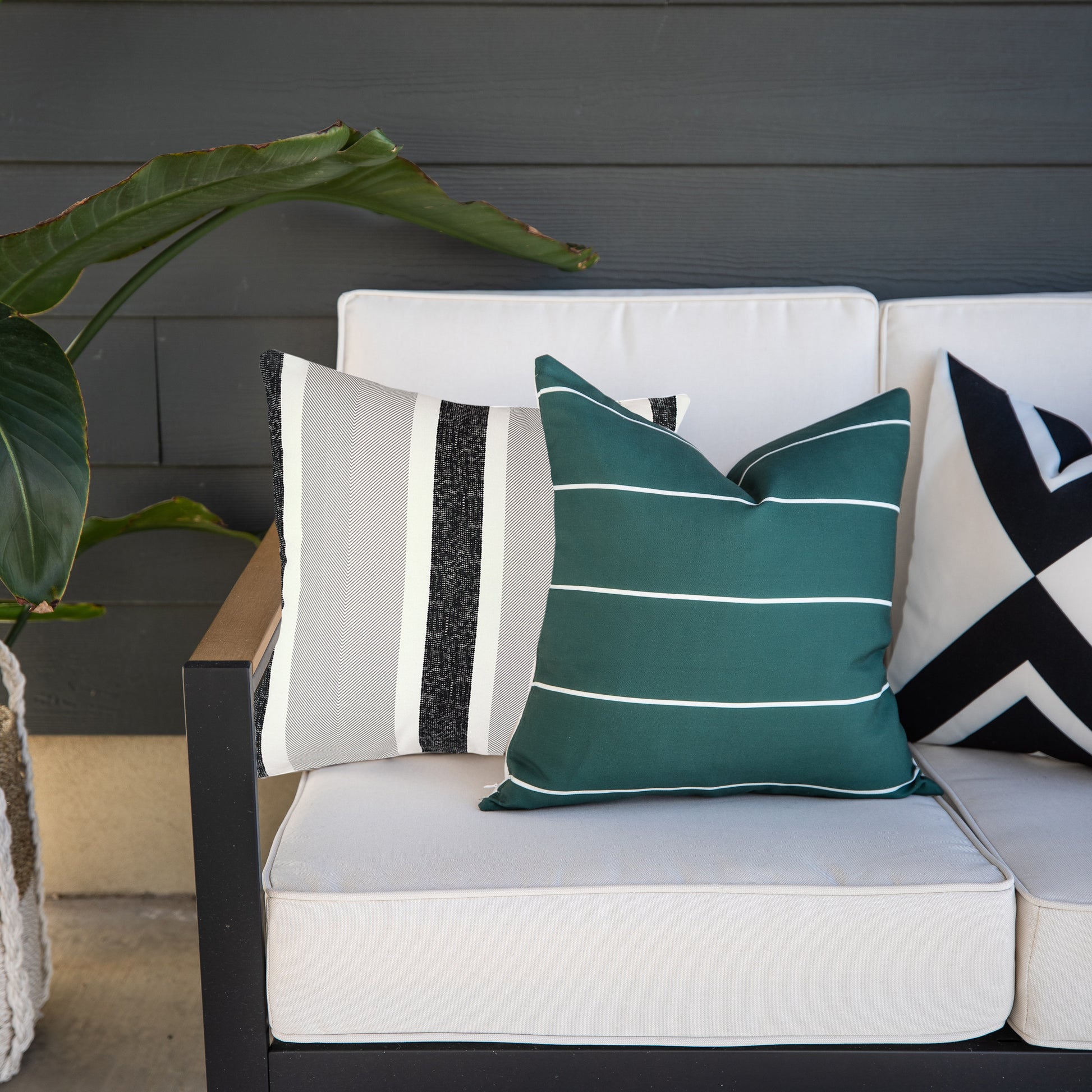 Four outdoor pillow covers displayed on a white cushioned chair: striped cream and charcoal, solid teal with white lines, and