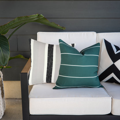 Four outdoor pillow covers displayed on a white cushioned chair: striped cream and charcoal, solid teal with white lines, and
