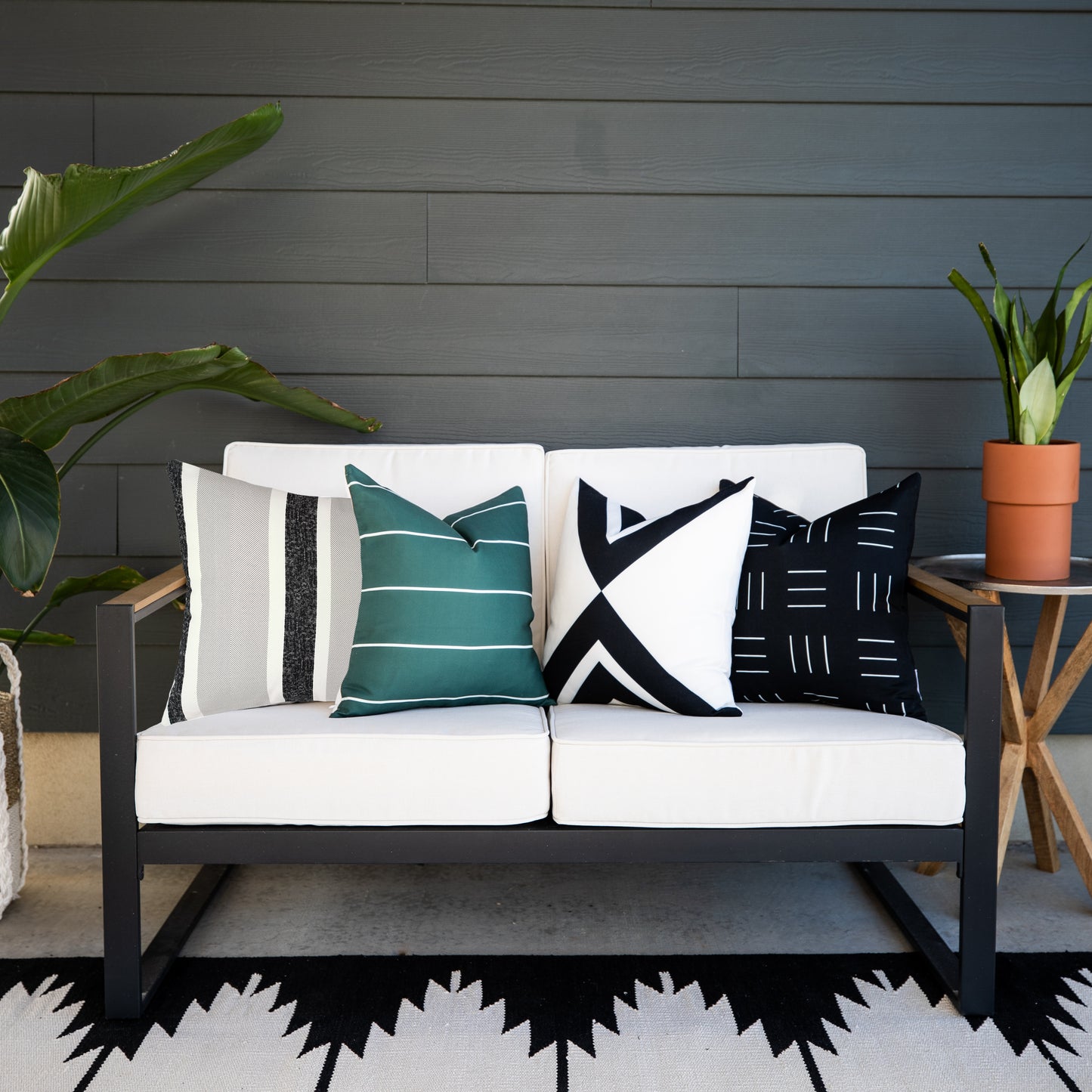 Four outdoor pillow covers in teal stripe, black geometric, cream and charcoal, and black dash patterns displayed on a black 