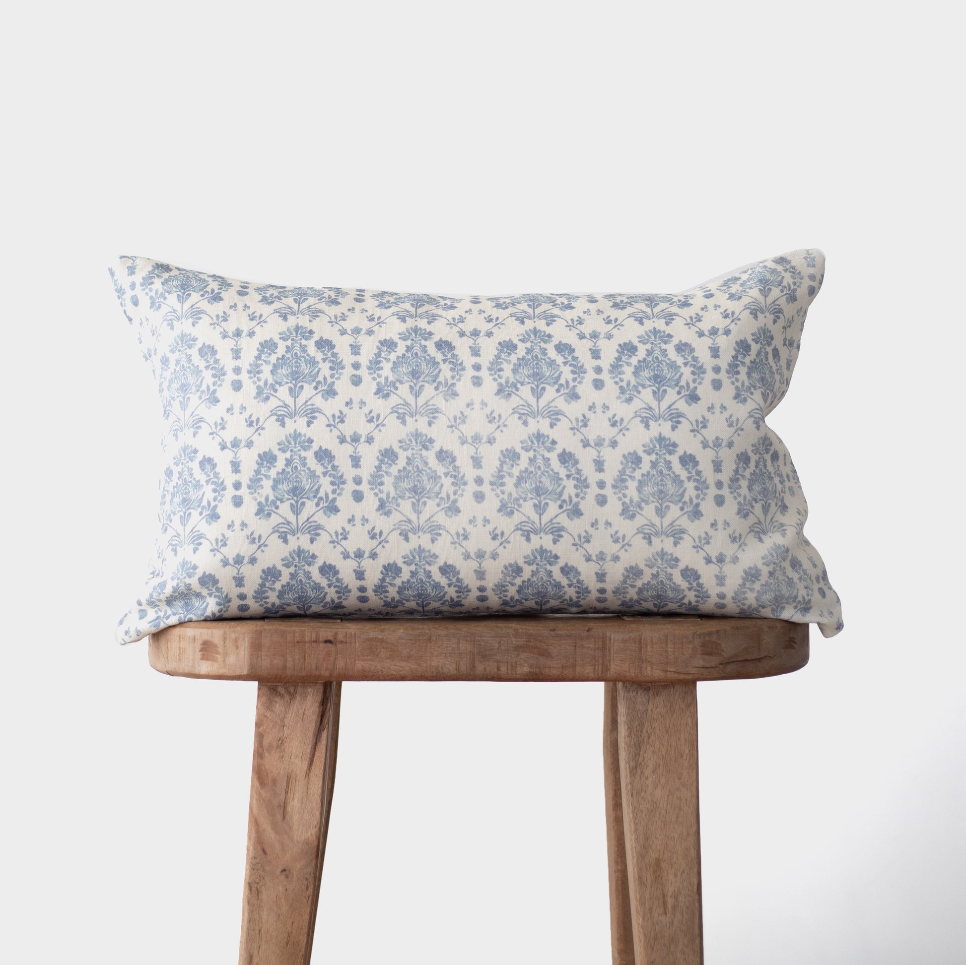 Blue and white damask patterned linen pillow on wooden bench against white background.