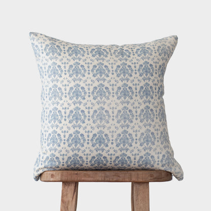 Blue and cream damask patterned linen pillow displayed on wooden chair back against gray background.
