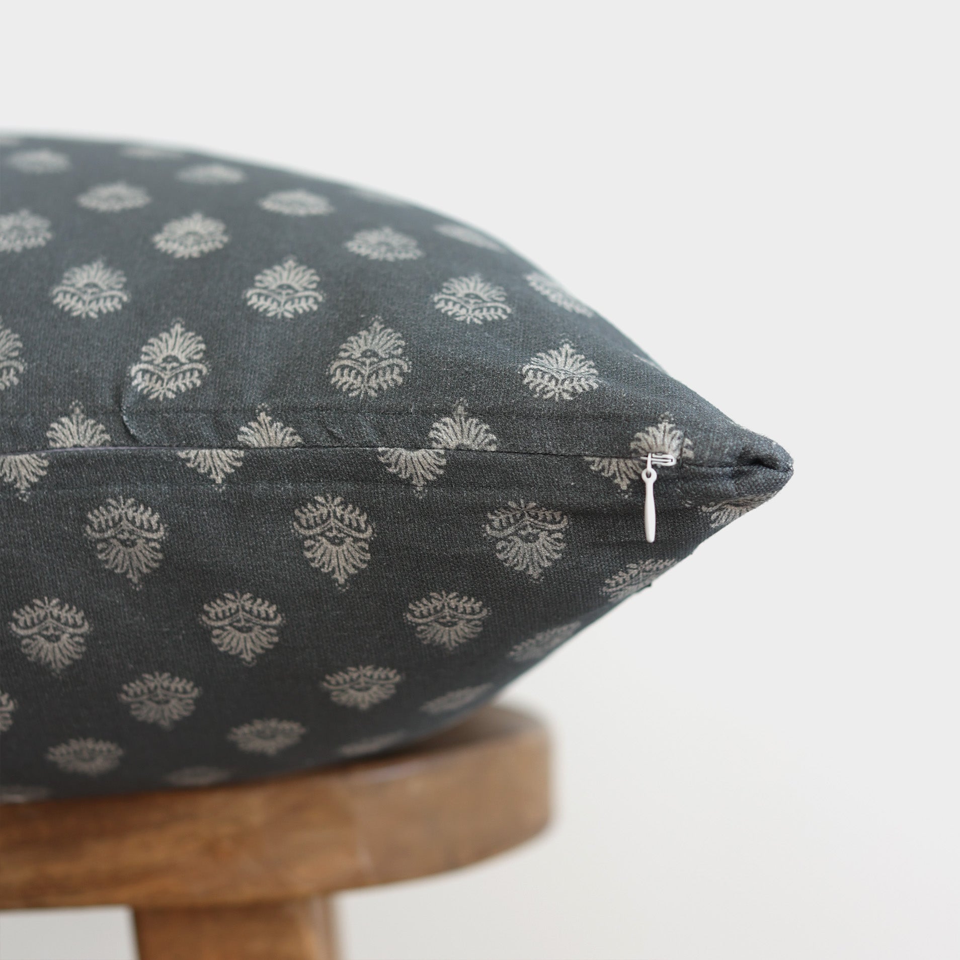 Blue-gray pillow with cream damask pattern and zipper detail resting on wooden stool.