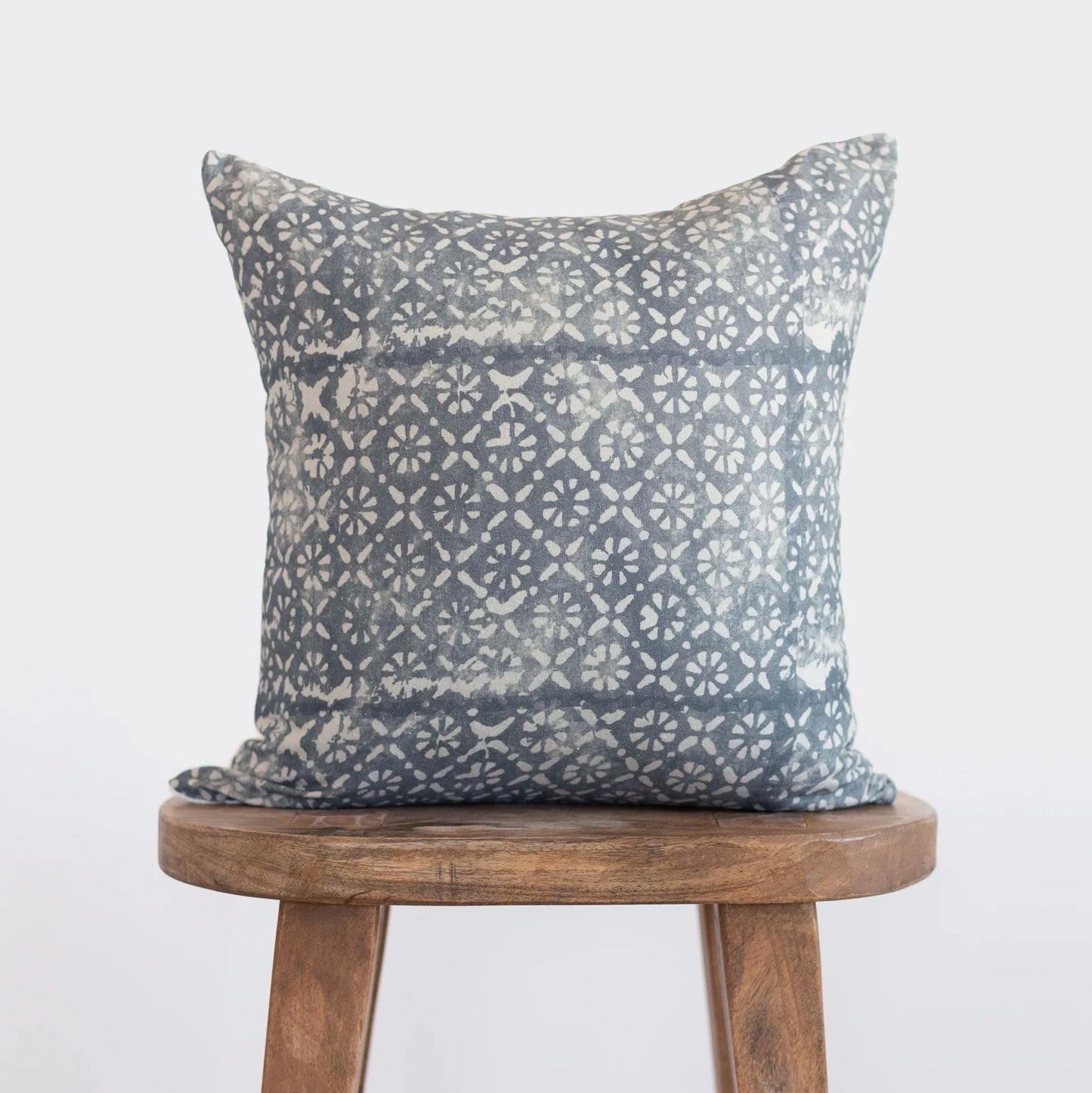 Blue-gray pillow with diamond and floral geometric pattern in cream, displayed on wooden stool.