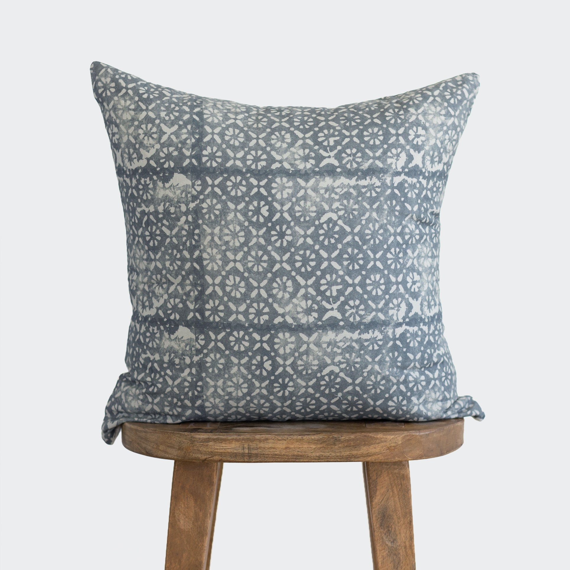 Blue-gray pillow with geometric diamond and floral pattern in cream, displayed on wooden stool.