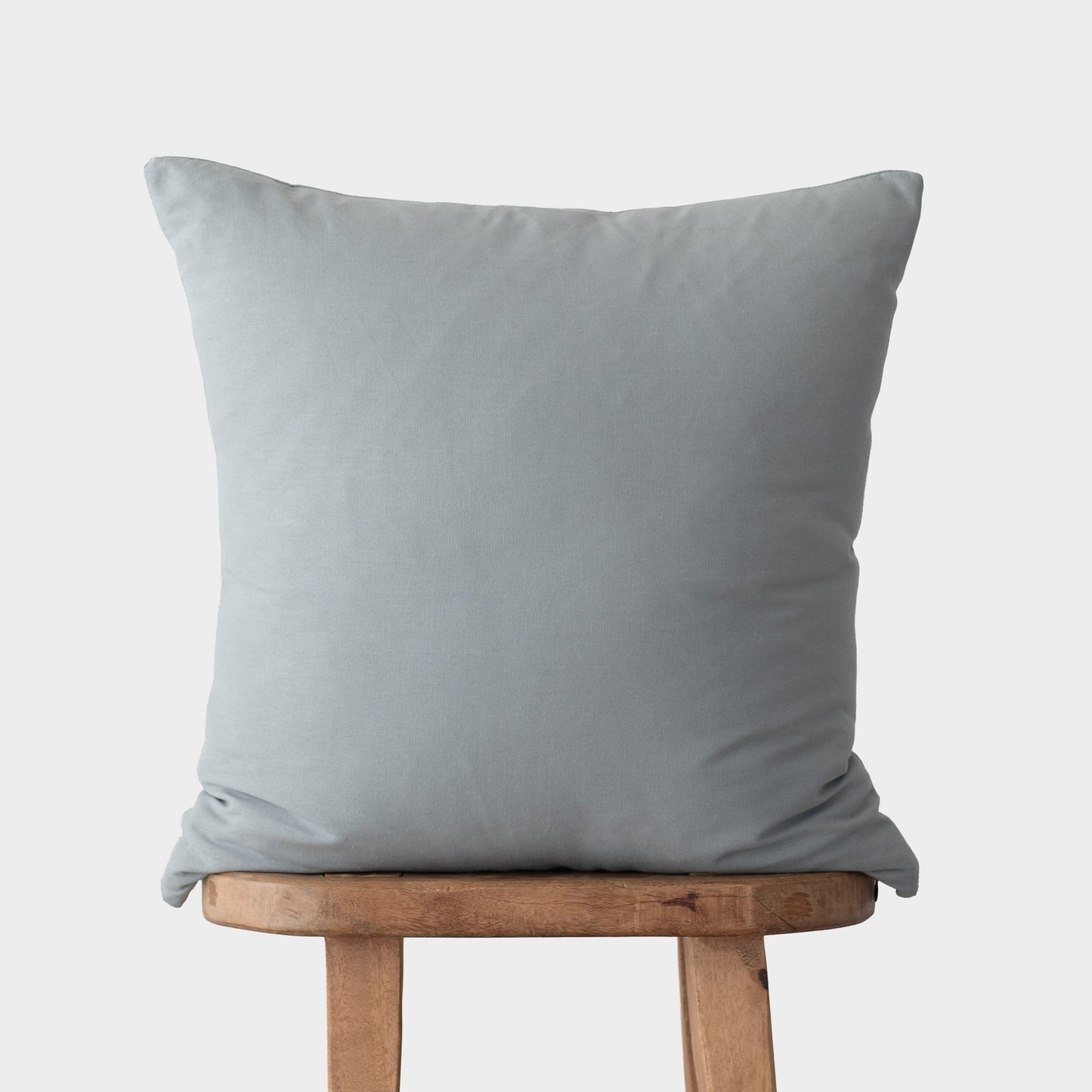 Dusty blue square throw pillow with knife edges displayed on wooden stool.