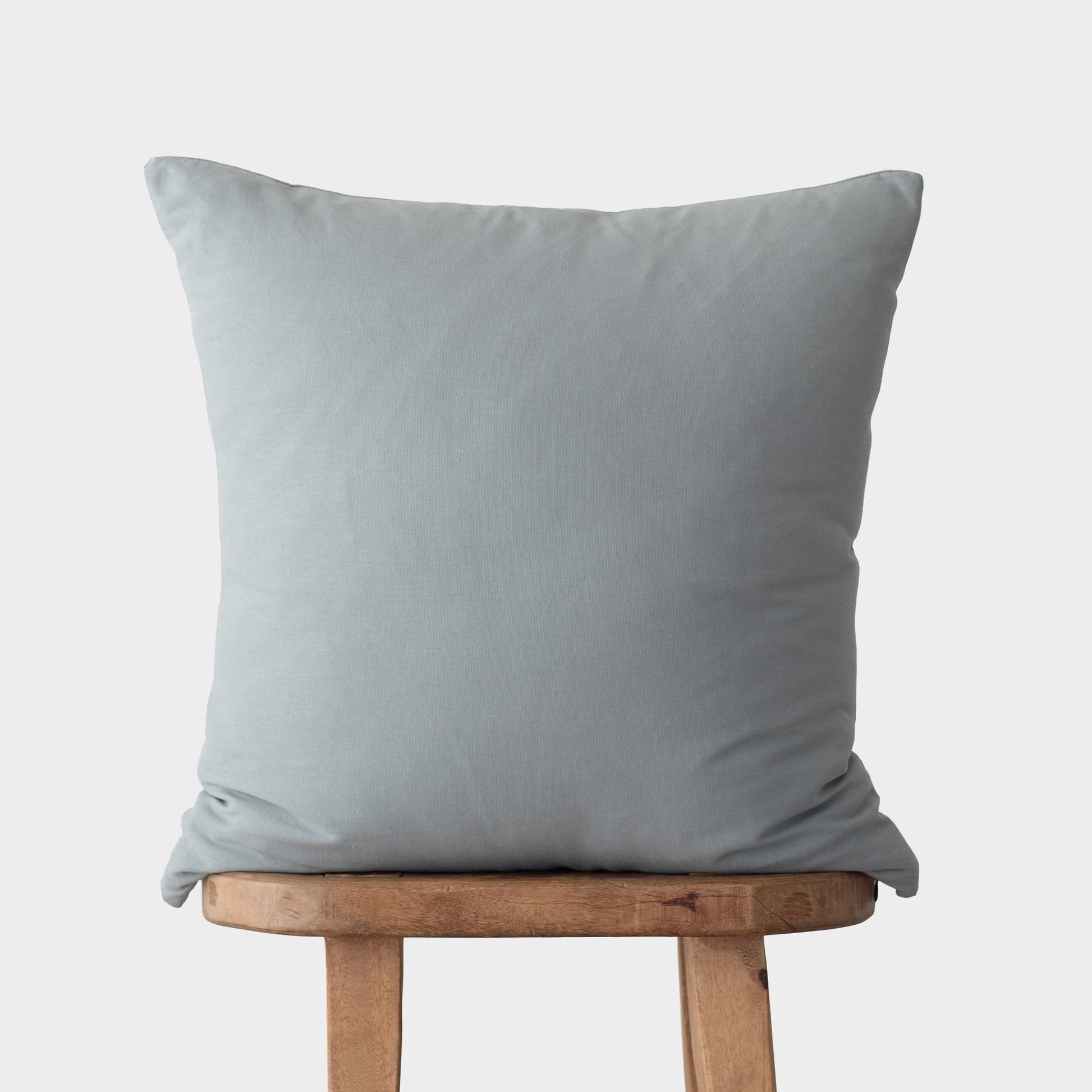 Dusty blue square throw pillow with knife edges displayed on wooden stool.
