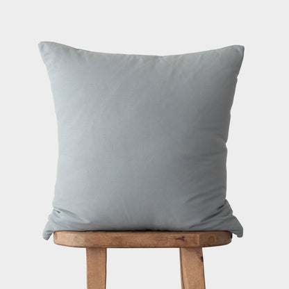 Dusty blue square throw pillow with knife edges displayed on wooden stool.