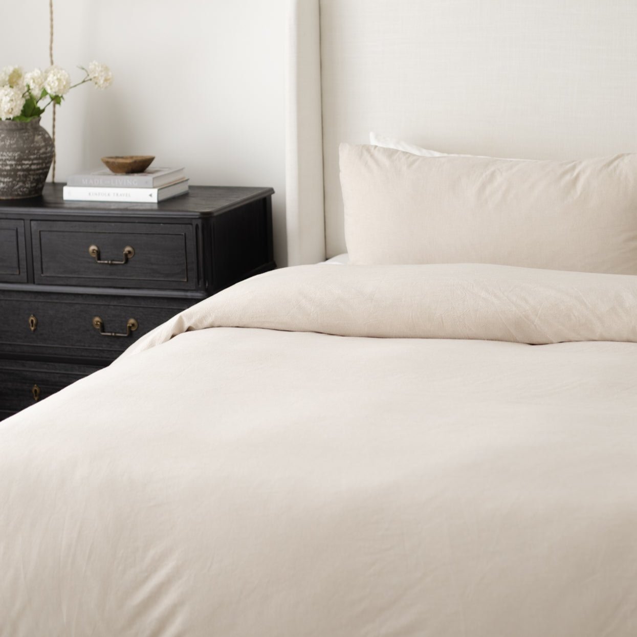 Beige duvet cover and pillowcases on a bed next to a black nightstand with white flowers and books.