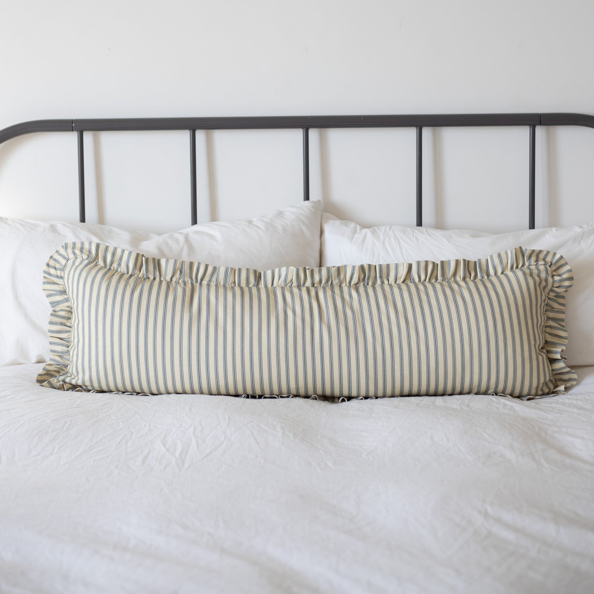 Long rectangular pillow with tan and white vertical stripes and ruffled edges on a bed with white bedding.