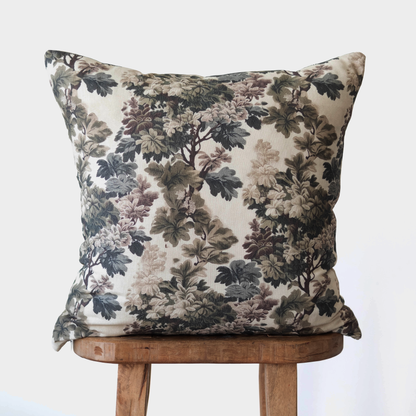 Square throw pillow with dark floral botanical print in black, gray, and mauve tones on cream background, displayed on wooden