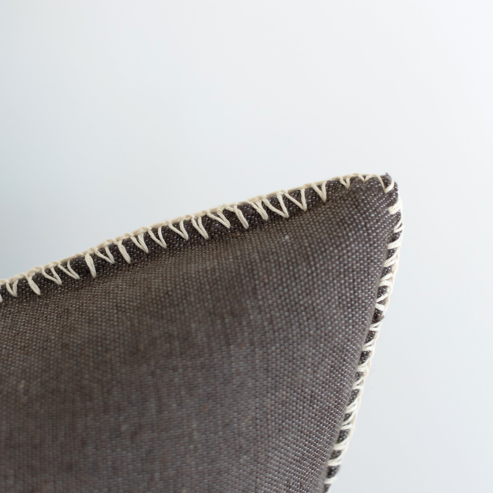 Warm charcoal woven pillow with cream whipstitched edges, shown at an angle.