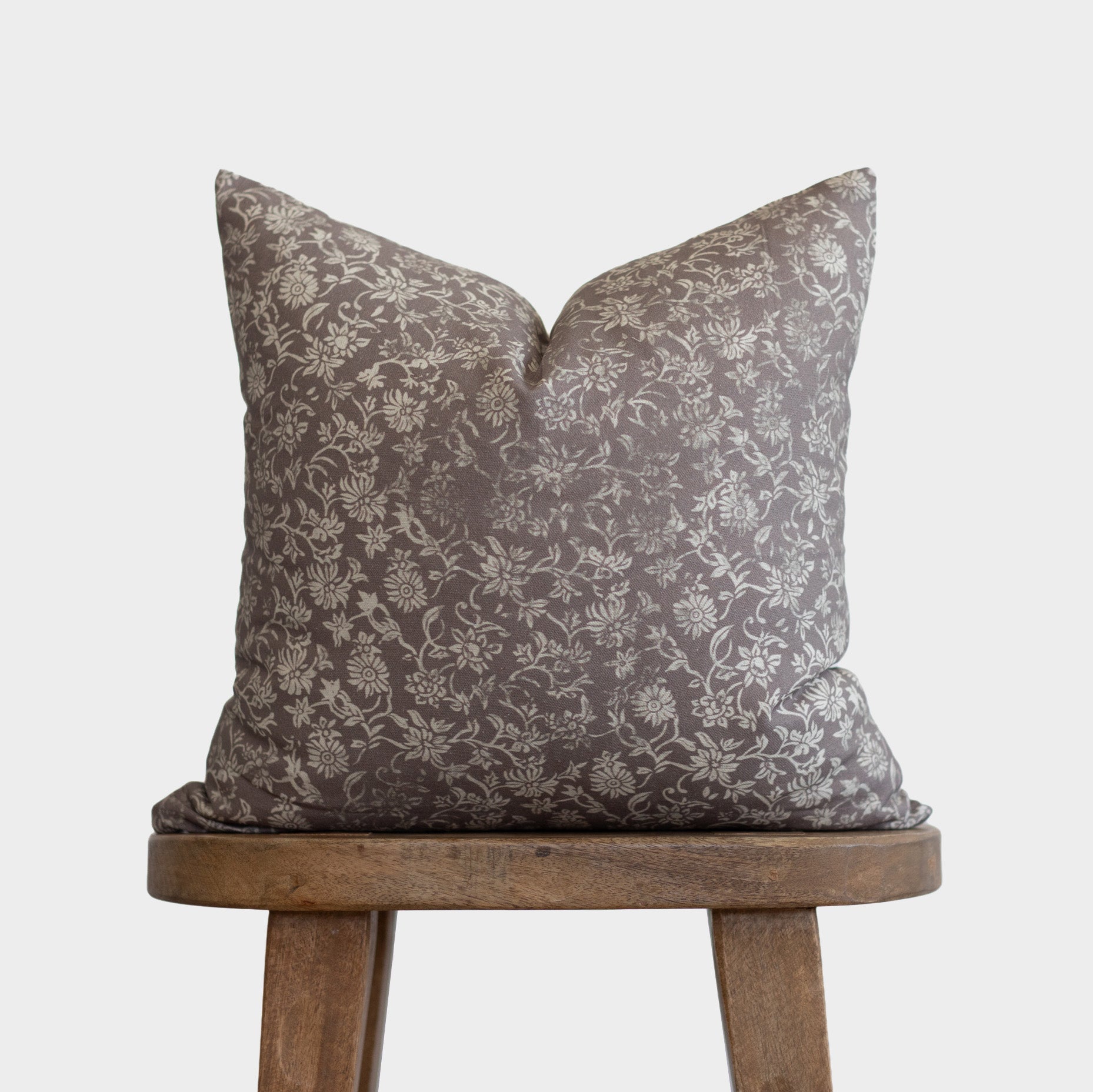 Gray and cream floral patterned pillow with envelope closure on wooden tripod stool.