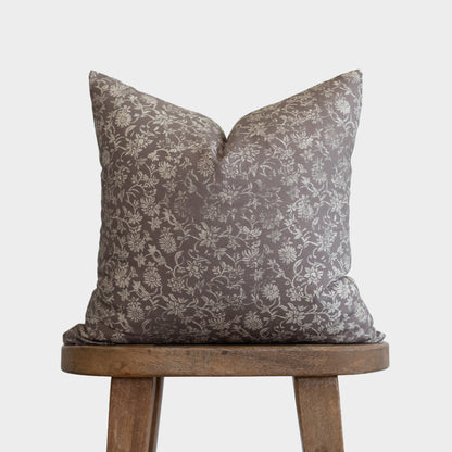 Gray and cream floral patterned pillow with envelope closure on wooden tripod stool.