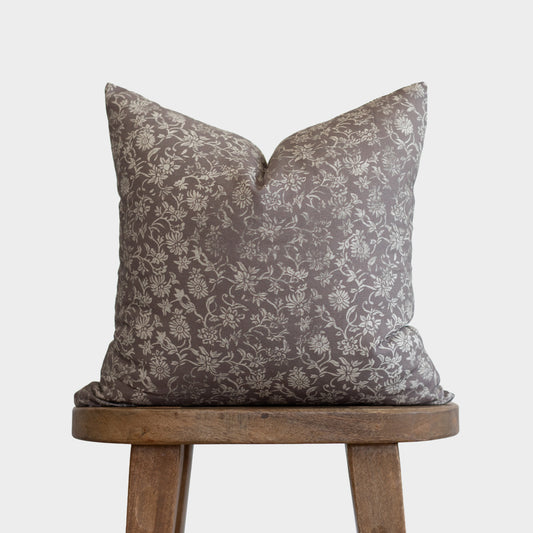 Gray and cream floral patterned pillow with envelope closure on wooden tripod stool.