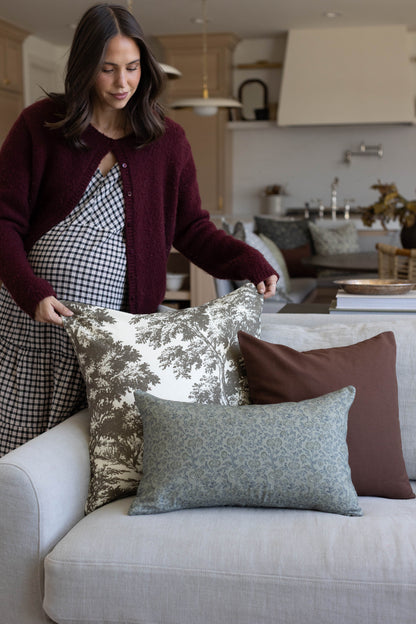 Woman arranging green and cream toile, solid brown, and patterned pillows on a light gray sofa.