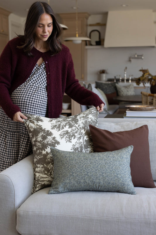 Woman arranging green and cream toile, solid brown, and patterned pillows on a light gray sofa.
