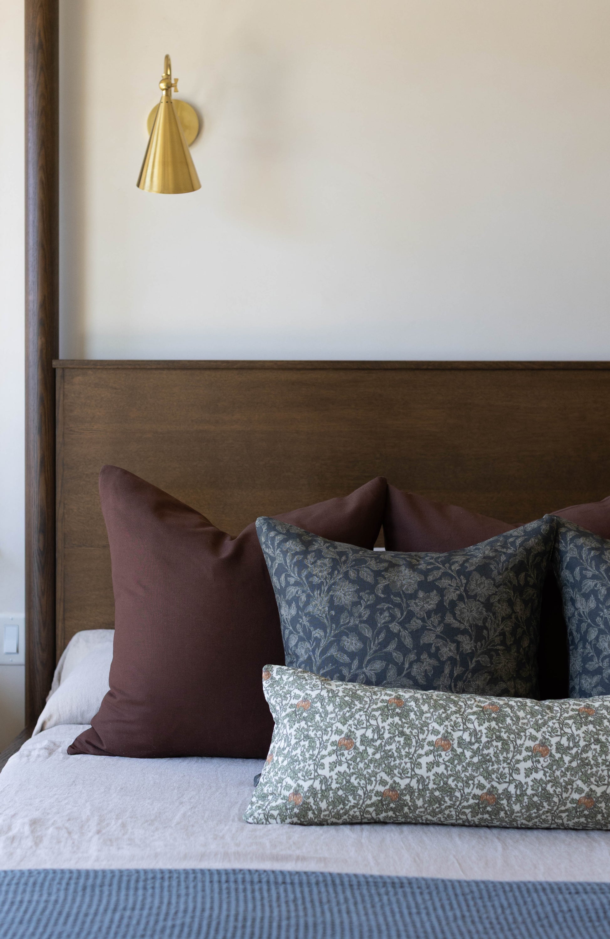 Brass cone-shaped wall sconce with finial top mounted above bed with burgundy and patterned pillows.
