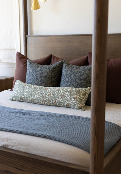Wooden four-poster bed styled with burgundy, patterned blue, and floral green pillows on white bedding.