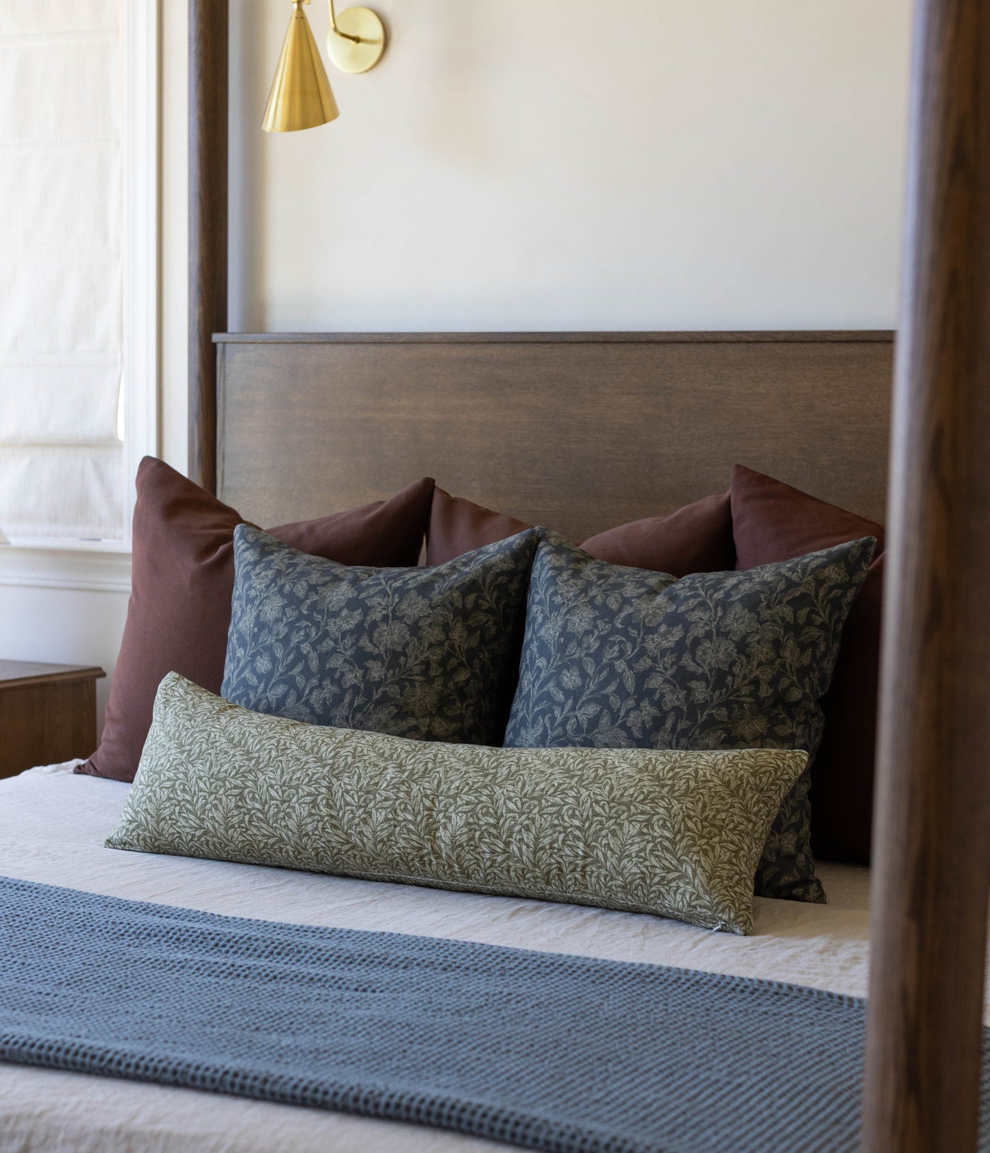 Green patterned lumbar pillow displayed on bed with burgundy and blue patterned pillows against gray headboard.
