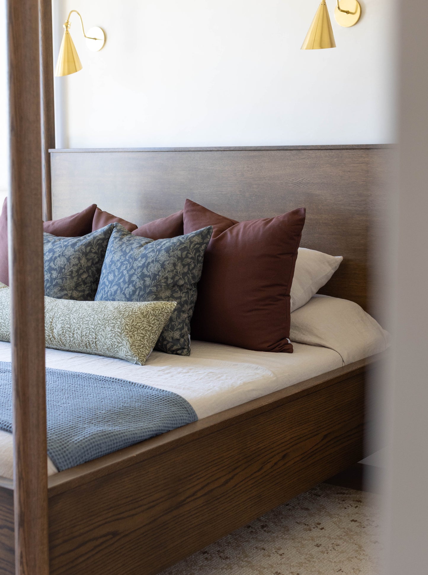 Bedroom with wooden bed frame, upholstered headboard, layered pillows in blue and burgundy tones, and brass wall sconces abov