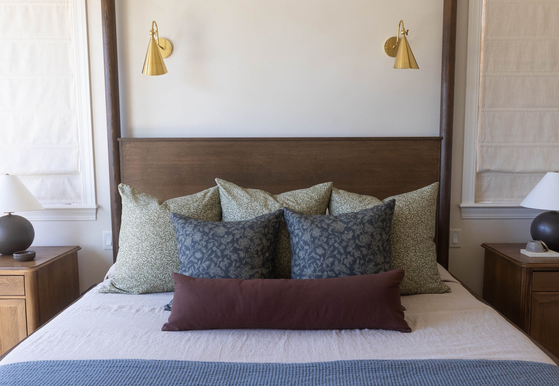 Bed with brown wooden headboard, patterned throw pillows in green and blue tones, burgundy lumbar pillow, and brass wall scon