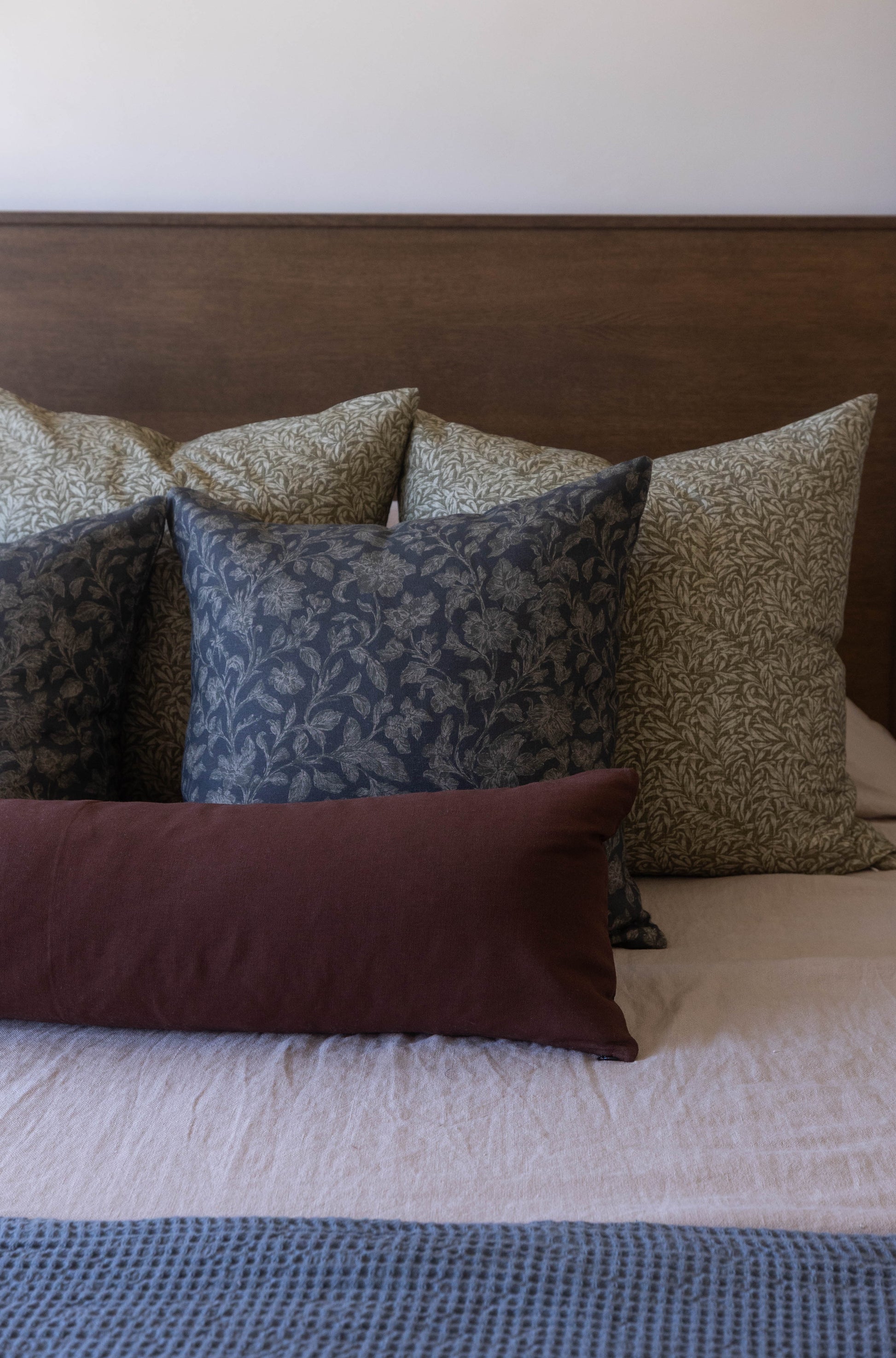Decorative pillows in navy, tan, and burgundy patterns arranged on a bed with brown headboard.