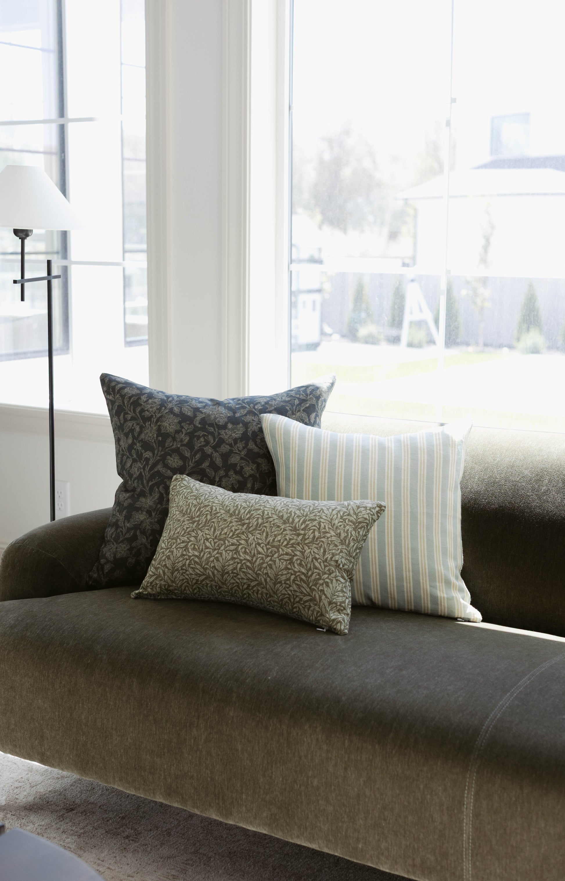 Three decorative pillows with damask, striped, and leaf patterns arranged on a gray upholstered sofa by a window.