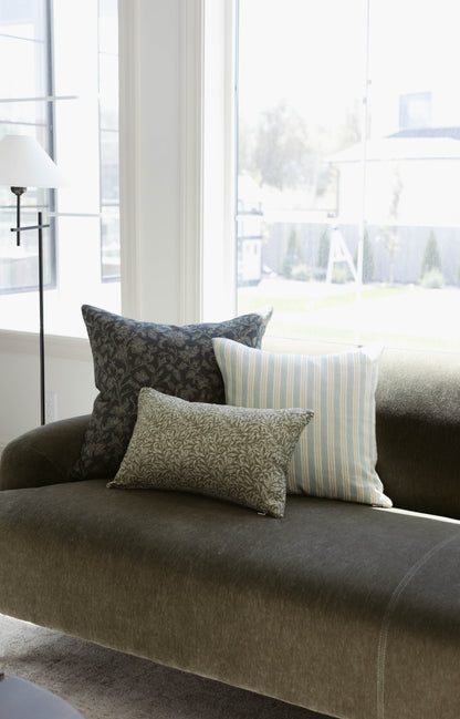 Three decorative pillows with damask, striped, and leaf patterns arranged on a gray upholstered sofa by a window.