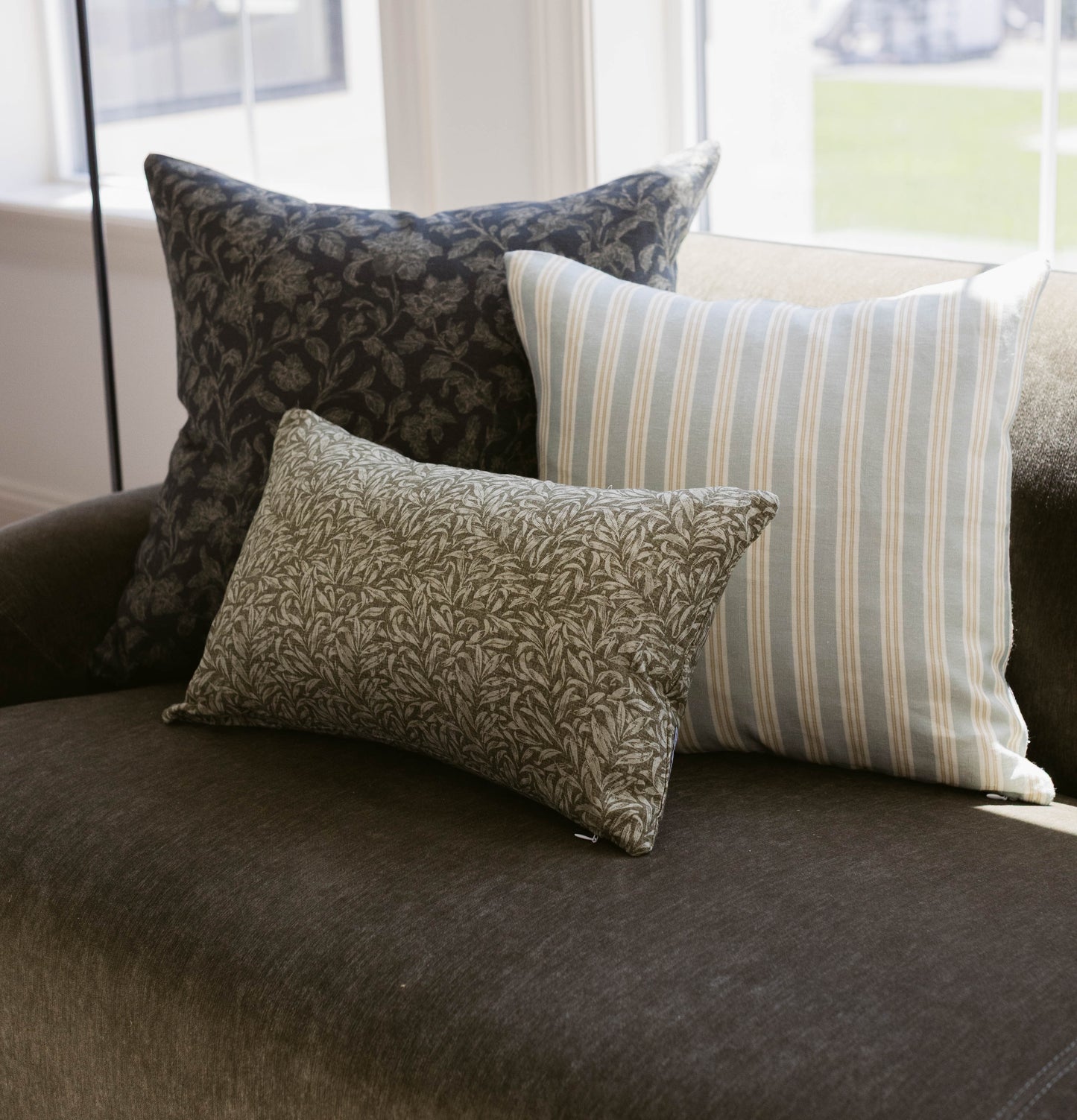 Three decorative pillows in damask, striped, and leaf patterns arranged on a dark upholstered chair by a window.