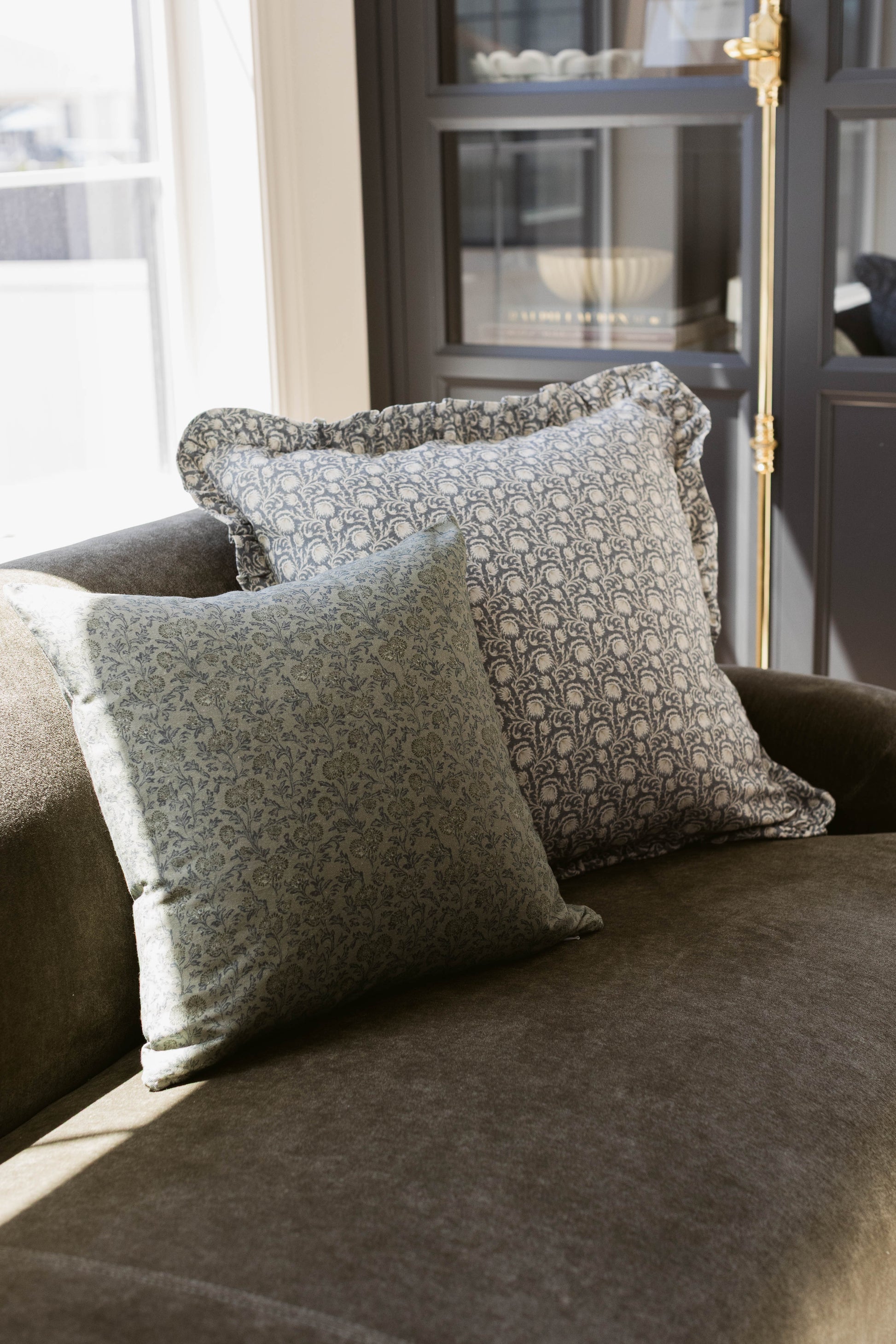 Two square decorative pillows with ruffled edges and damask pattern on gray upholstered chair.