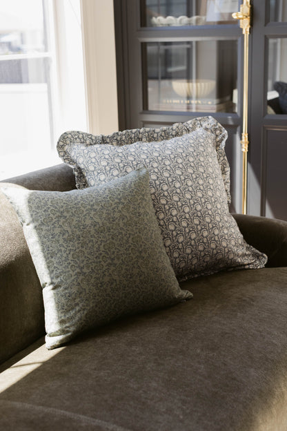 Two square decorative pillows with ruffled edges and damask pattern on gray upholstered chair.