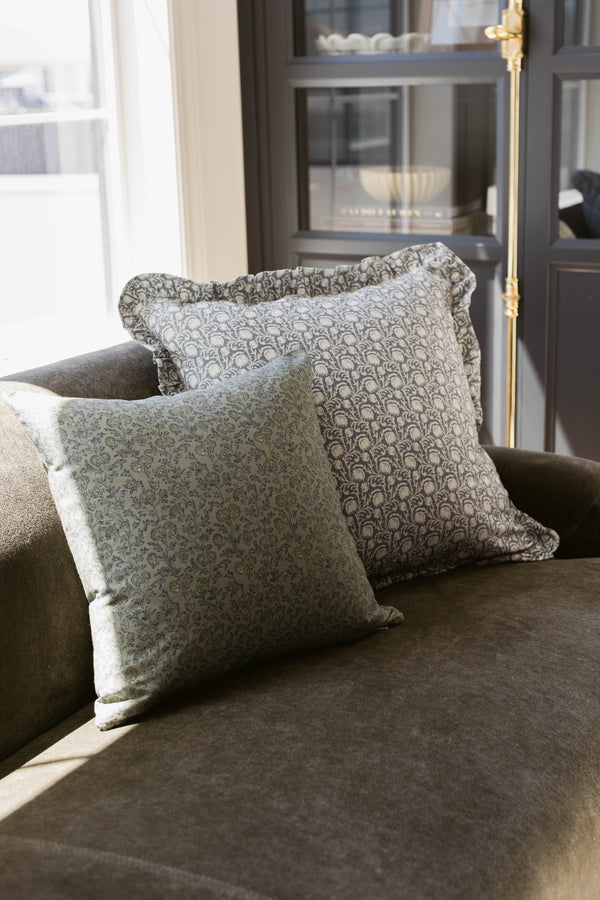 Two square decorative pillows with ruffled edges and damask pattern on gray upholstered chair.