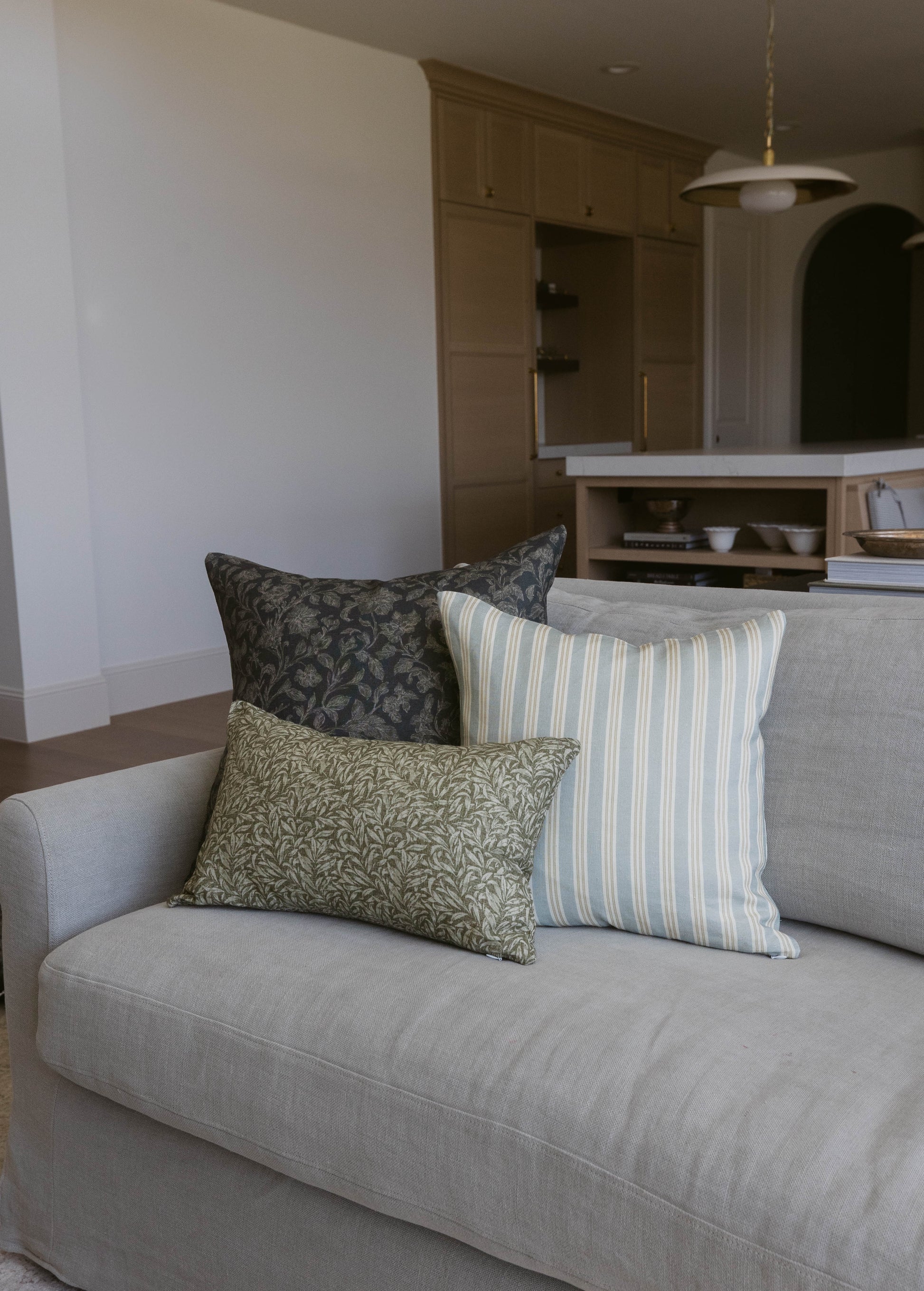 Three decorative pillows on a gray upholstered sofa: dark patterned, green botanical, and blue striped designs.