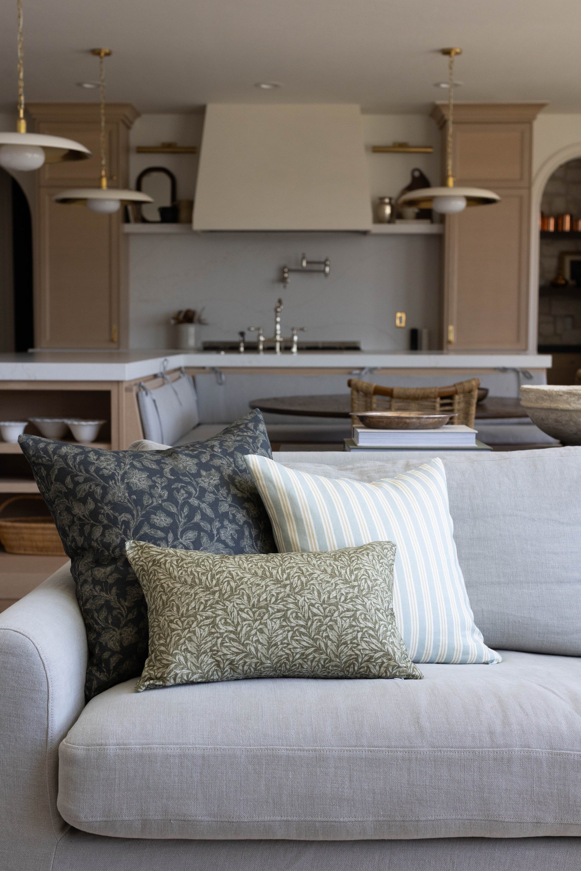 Three decorative pillows on a light gray sofa in shades of navy, cream, and sage green with patterned and striped designs.
