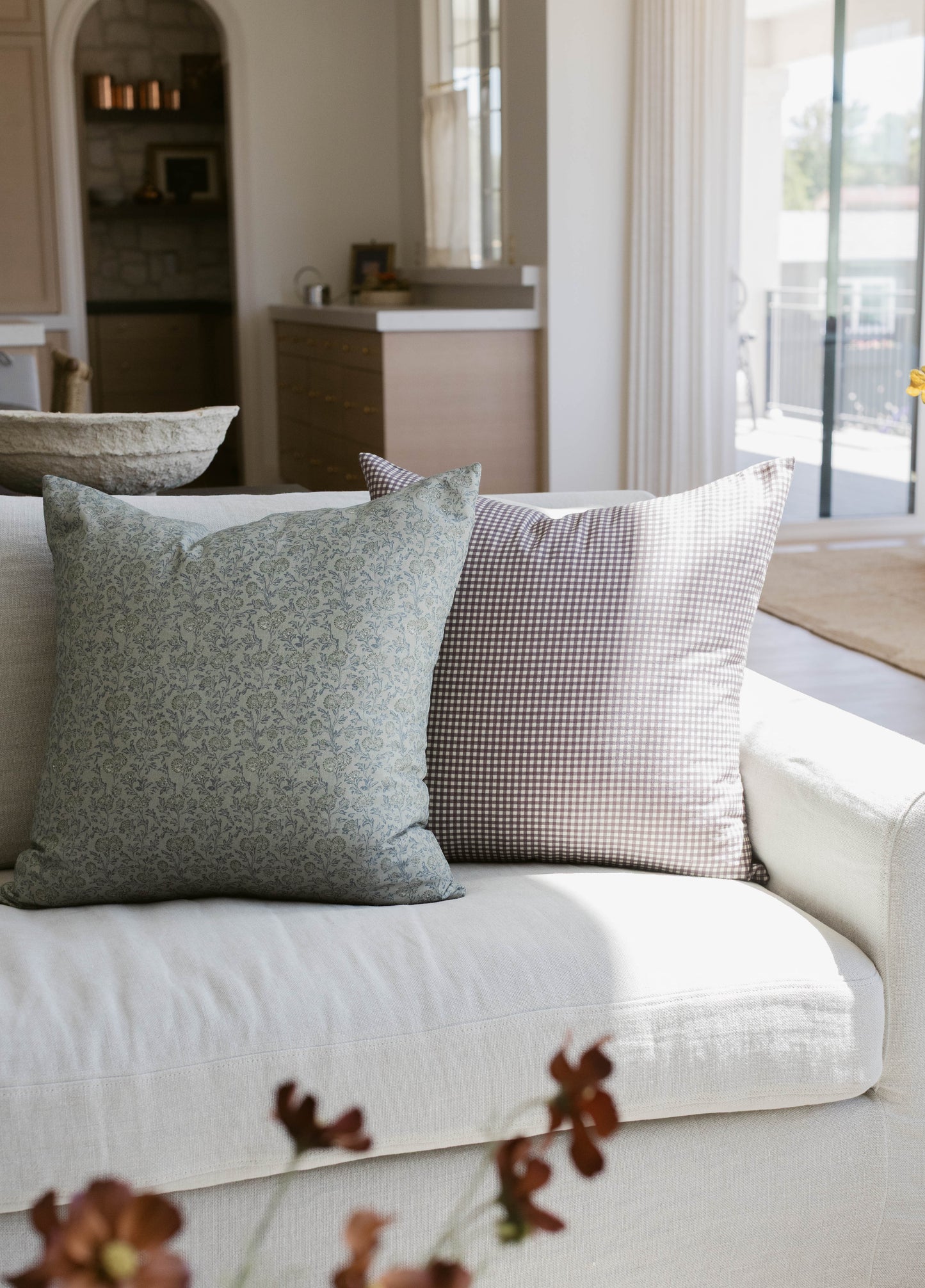 Two decorative pillows on a cream sofa: one sage green with floral pattern, one brown and white gingham check.
