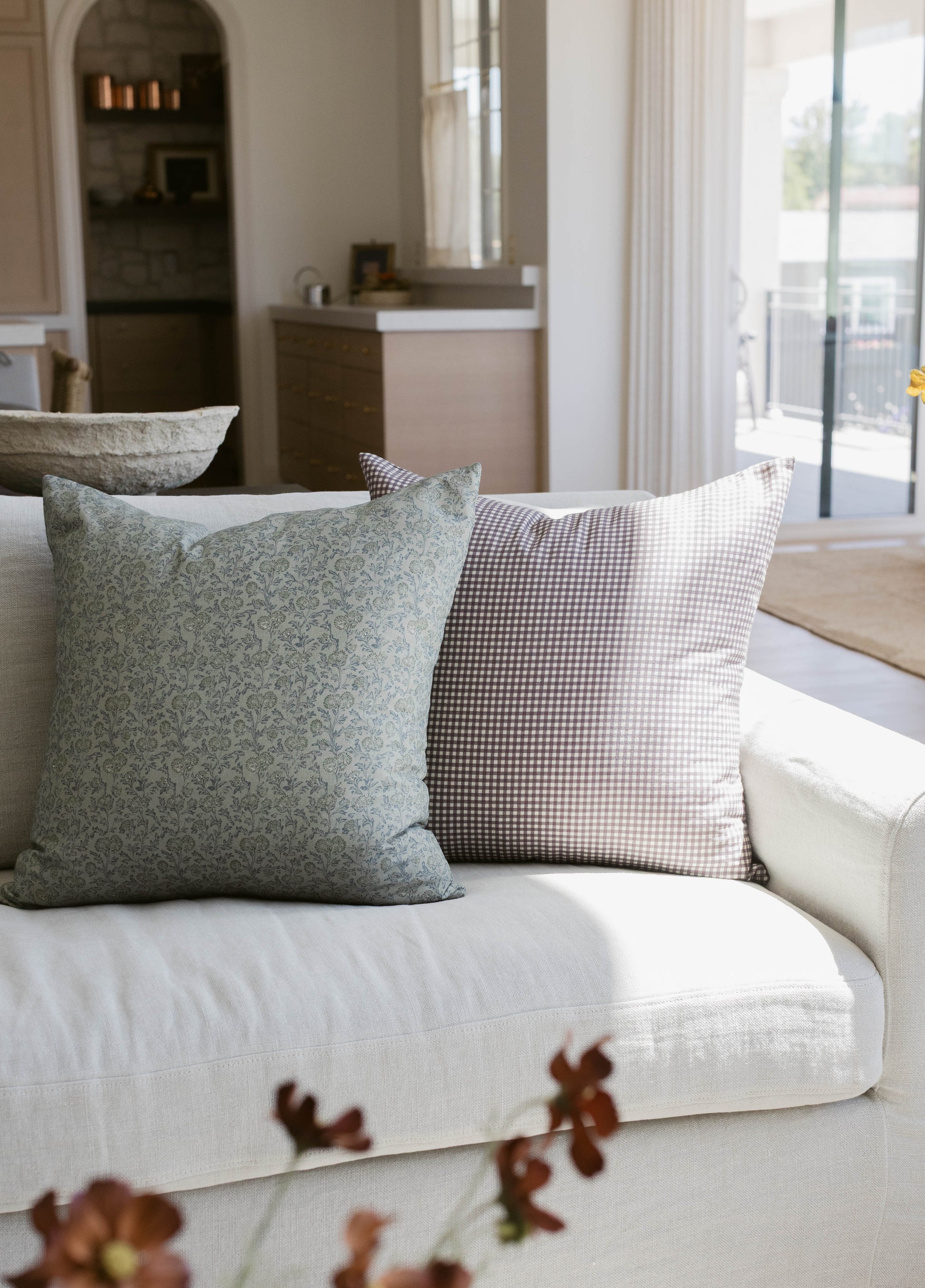 Two decorative pillows on a cream sofa: one sage green with floral pattern, one brown and white gingham check.