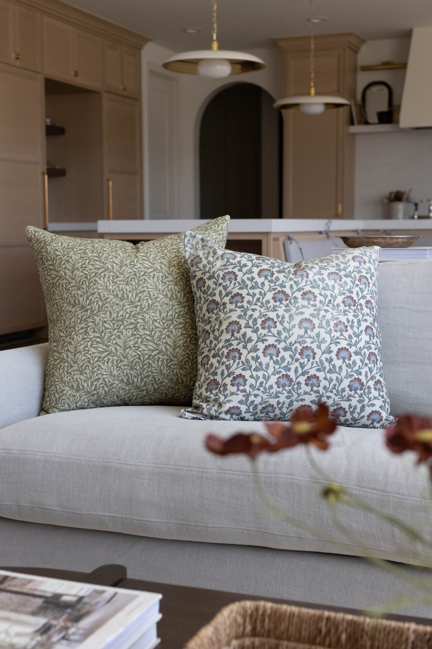 Two multicolor patterned throw pillows with floral and leaf designs in sage and blue tones on a cream sofa.