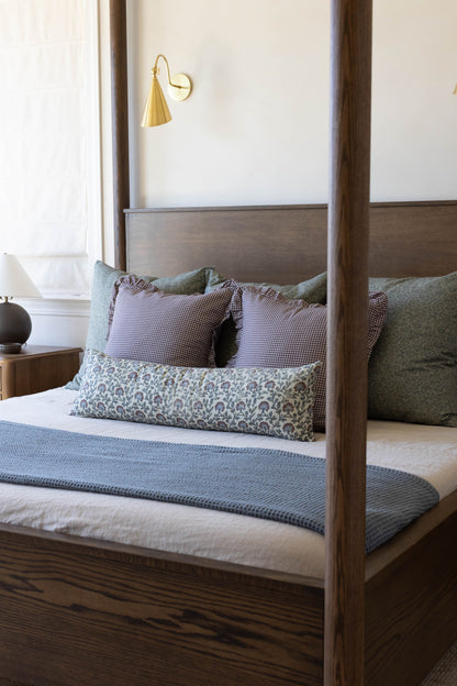 Gold cone-shaped wall sconce with curved arm mounted above wooden bed with multicolor pillows.