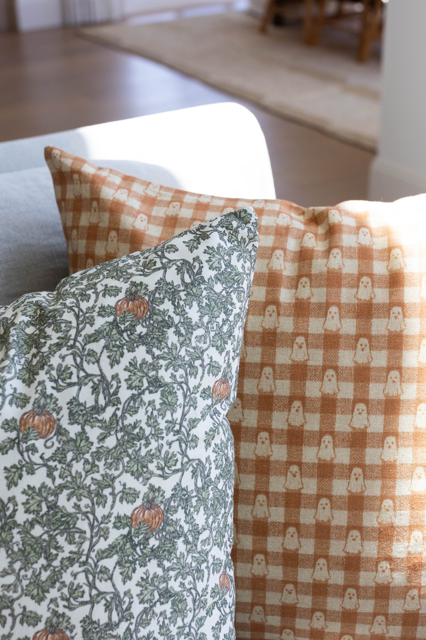 Tan and cream gingham pillow with ghost pattern, displayed next to floral patterned pillow on chair.