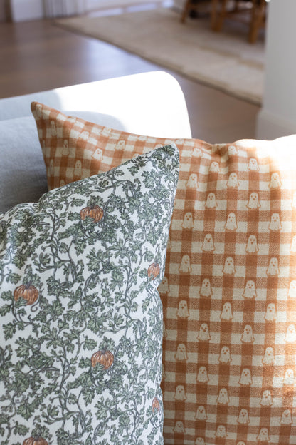 Tan and cream gingham pillow with ghost pattern, displayed next to floral patterned pillow on chair.
