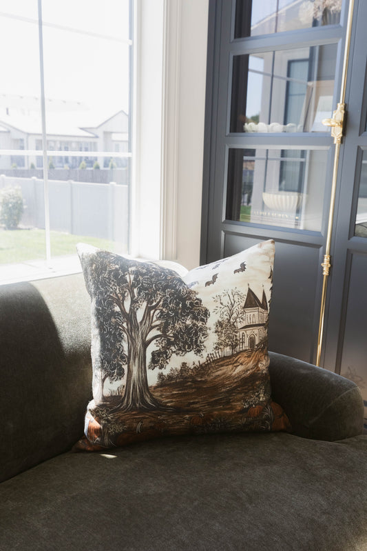 Linen pillow featuring a haunted house, gnarled tree, and flying bats in black and brown tones on cream background.